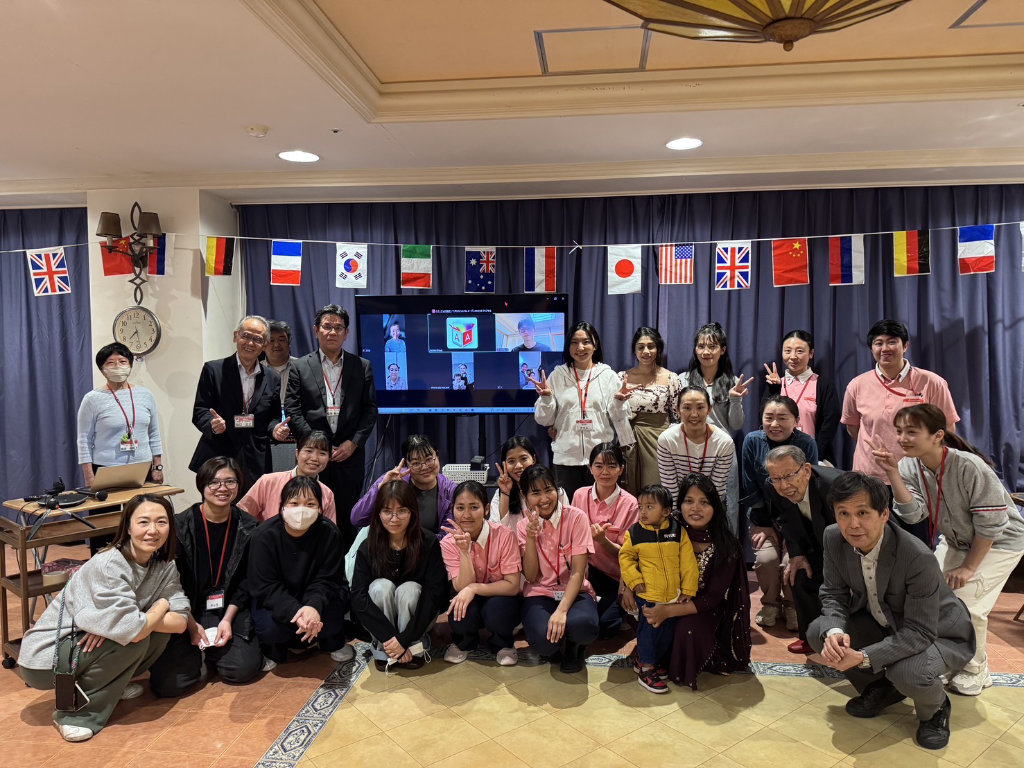 Foreign staff and Japanese staff and management posing for a photo in front of a screen showing a virtual attendees, surrounded by international flags