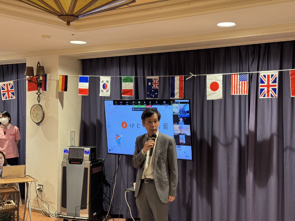 Dr Koji Kurusu delivering opening remarks with a microphone in front of a presentation screen, flanked by international flags