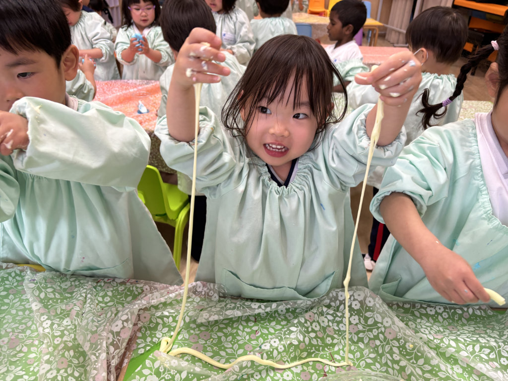 Children engage in a hands-on STEAM activity, smiling and stretching gooey slime-like material at a classroom table, exploring textures through play.