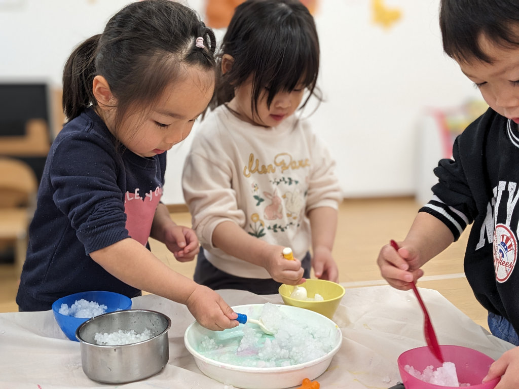 Three young children participate in a STEAM activity, using spoons and colorful bowls to explore icy materials, focused and engaged in sensory learning.