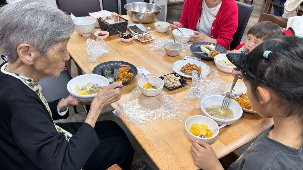 An elderly woman, two children, and an adult gather around a table, sharing a meal with a variety of dishes in small bowls and plates. The table includes soup, fruit, rice, and vegetables. The woman holds a bowl of food, while the children eat attentively.