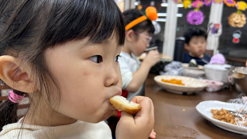 A young girl eats a piece of bread, while two other children sit at the table with food. Halloween decorations hang in the background.