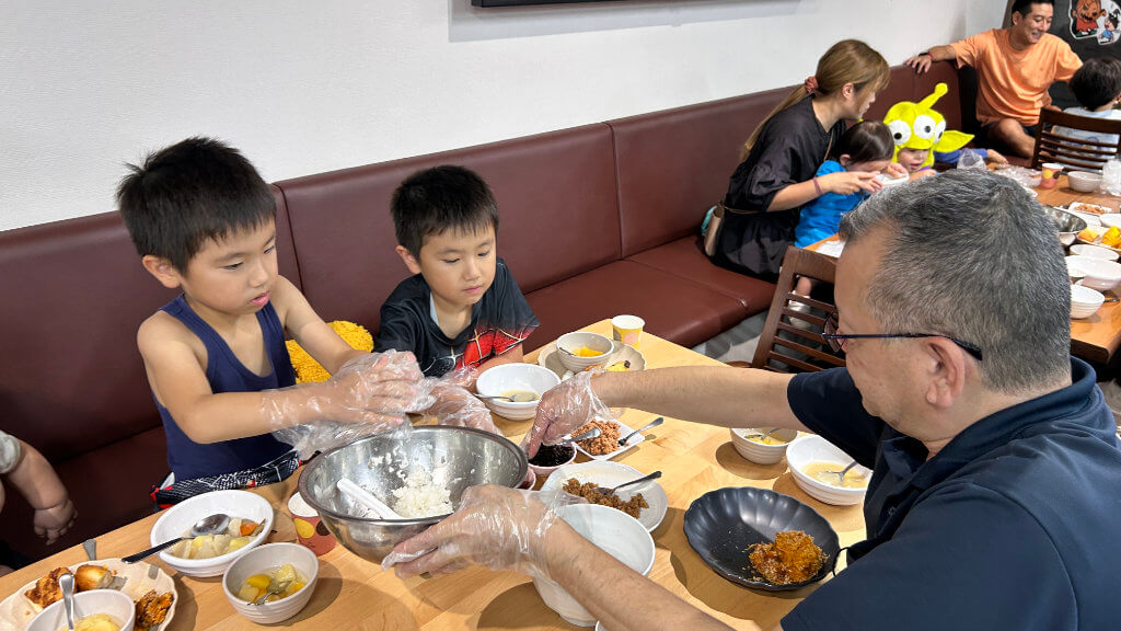 Two young boys and an adult with gloves mix food in a bowl at a table, sharing a meal. More families dine in the background.