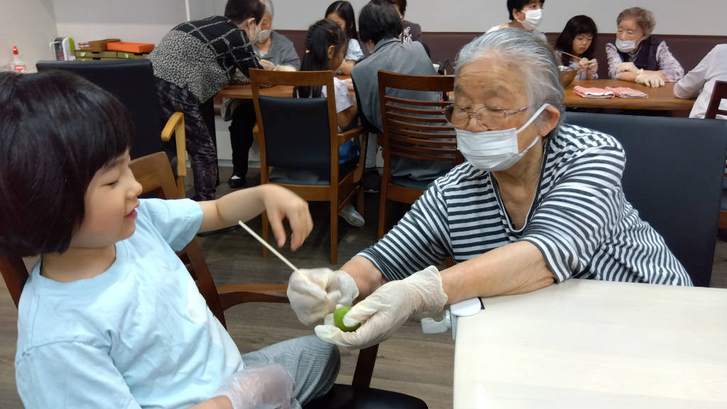 An elderly woman and a child are seated at a table, both wearing gloves and masks, working together at removing a plum stem